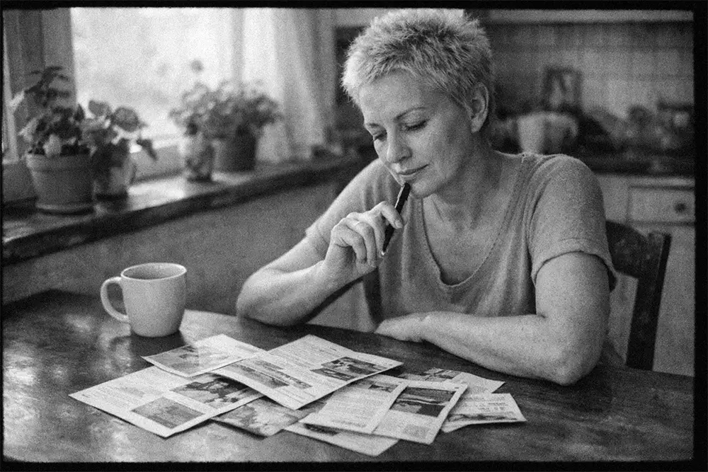 Femme d’une quarantaine d’années assise à une table de cuisine, lisant des brochures avec un stylo à la main, ambiance calme en noir et blanc style photo argentique