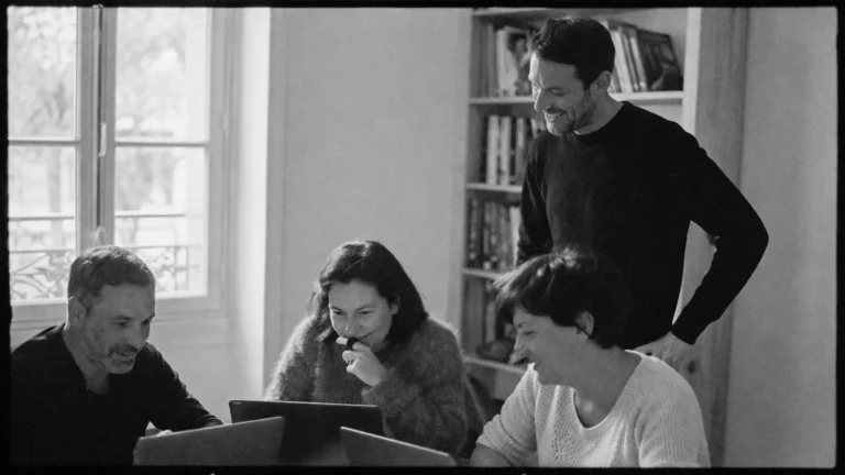 Petit groupe d’adultes en formation autour d’une table avec ordinateurs portables, accompagné par un formateur debout, ambiance naturelle et collaborative en noir et blanc
